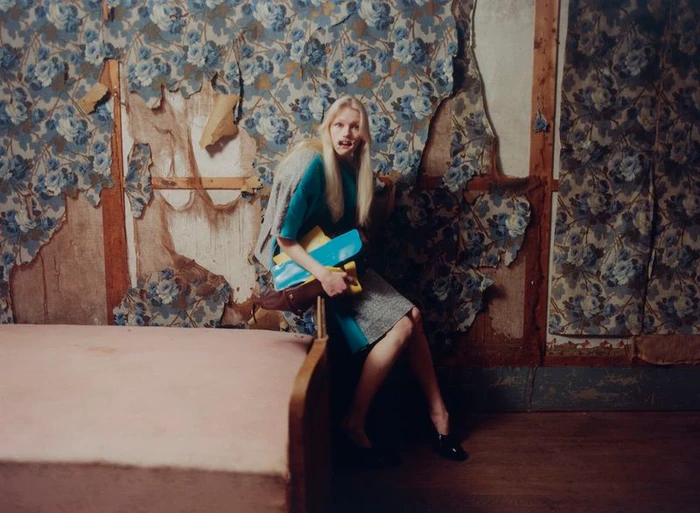 Model with long blonde hair holding books and bags seated in a derelict room with peeling floral wallpaper