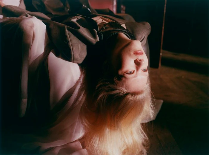 Model Malin Rudnick lying inverted with blonde hair draped downwards, wearing an oversized jacket and layered fabric.