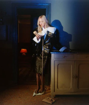 Model Malin Rudnick in a black patent coat with white collar accents holding a landline phone receiver beside a wooden cabinet.