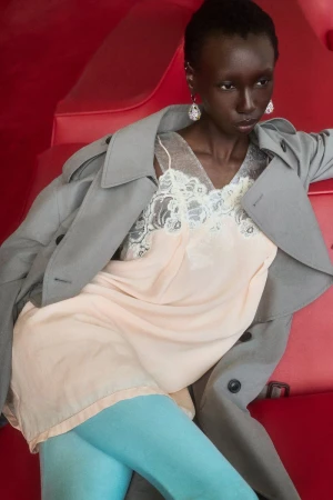 Model Alay Deng reclining on red seating in a pale slip dress, grey trench coat, and light blue tights.
