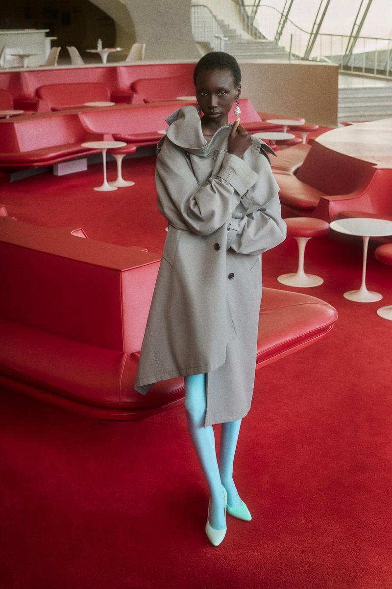 Model in a grey coat and bright blue tights standing inside a lounge with red seating and carpeting for WWD.