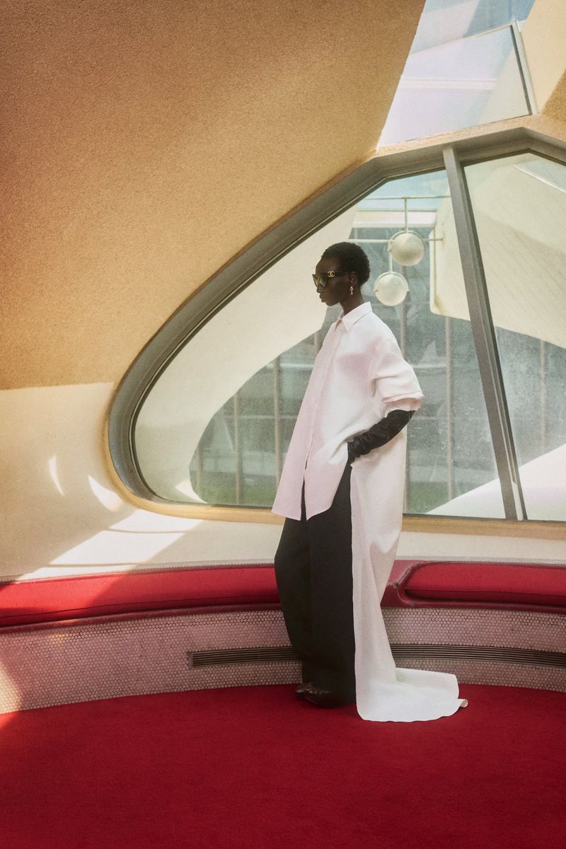 Alay Deng wearing an oversized white trailing tunic, black trousers, and sunglasses standing on red carpet by curved windows.