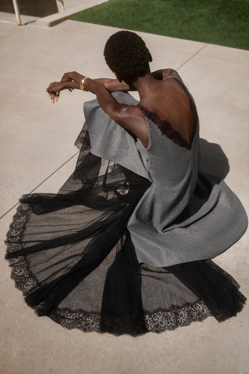 High-angle view of model Alay Deng seated on concrete wearing a grey dress with an expansive black lace trim skirt.