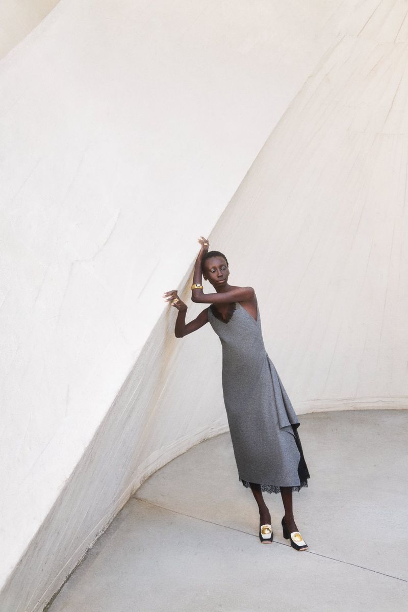 Model Alay Deng wearing a grey midi dress and gold jewelry, leaning against a large slanted white architectural wall.