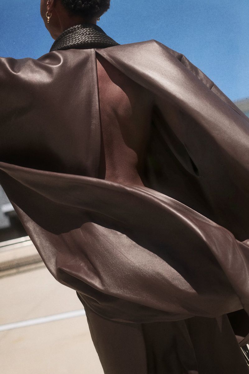 Model Alay Deng from behind wearing an open-back brown oversized leather coat against a blue sky.