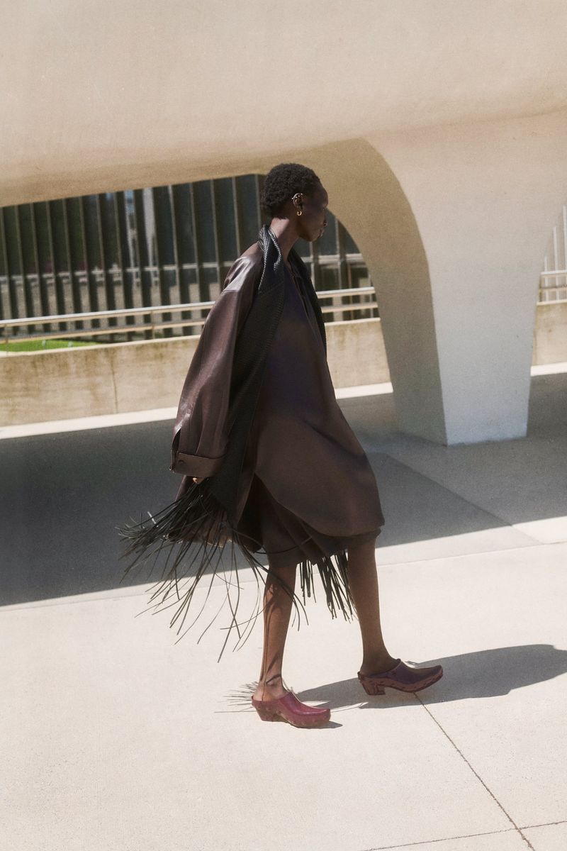 Model Alay Deng walking in a dark brown fringed coat and wooden clogs beside curved concrete architecture.