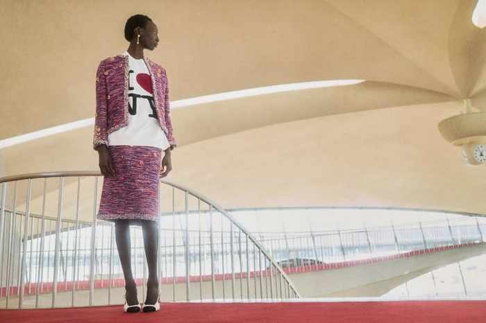 Model Alay Deng stands in a purple tweed skirt suit and I Love NY t-shirt inside an airport terminal with vaulted ceilings.