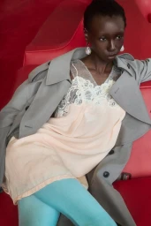 Model Alay Deng reclining on red seating in a pale slip dress, grey trench coat, and light blue tights.