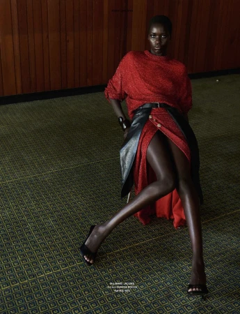 Model Awar Odhiang seated in a red knit dress with a black leather overlay on a patterned carpet against a wood-paneled wall.