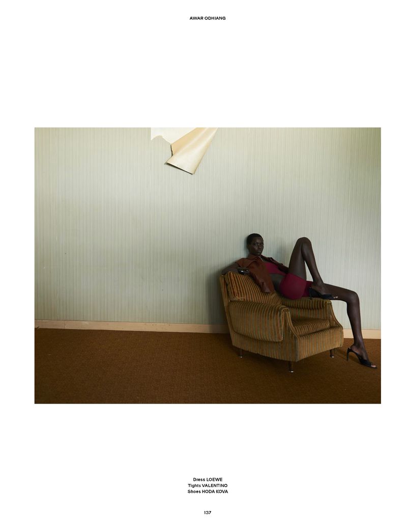 Model Awar Odhiang reclining on a corduroy armchair against a pale striped wall with peeling wallpaper in a magazine editorial.