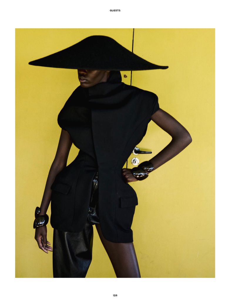 Model Awar Odhiang wearing a sculptural wide-brim black hat and structured black vest in front of a bright yellow door.