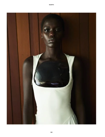Model Awar Odhiang wearing a white sleeveless dress with a glossy dark chest piece against a vertical wood paneled wall.