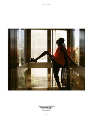 Model Awar Odhiang posing dynamically inside a glass elevator wearing an orange draped top and black boots in natural daylight.