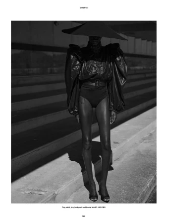 Model Awar Odhiang in an oversized sculptural black hat, voluminous jacket, and bodysuit posing on concrete stadium steps.
