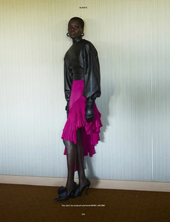 Model Awar Odhiang posing in a black leather top, vibrant magenta ruffled skirt, and black heels against a pale ribbed wall.