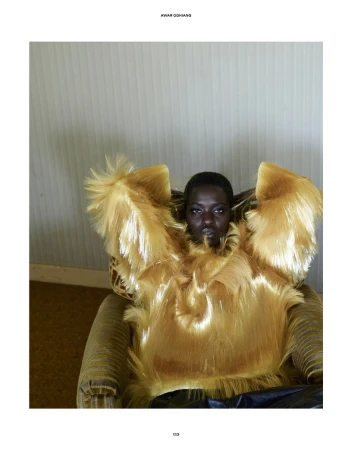 Model Awar Odhiang in a voluminous blonde faux-fur textured top seated in an armchair for The Travel Almanac.