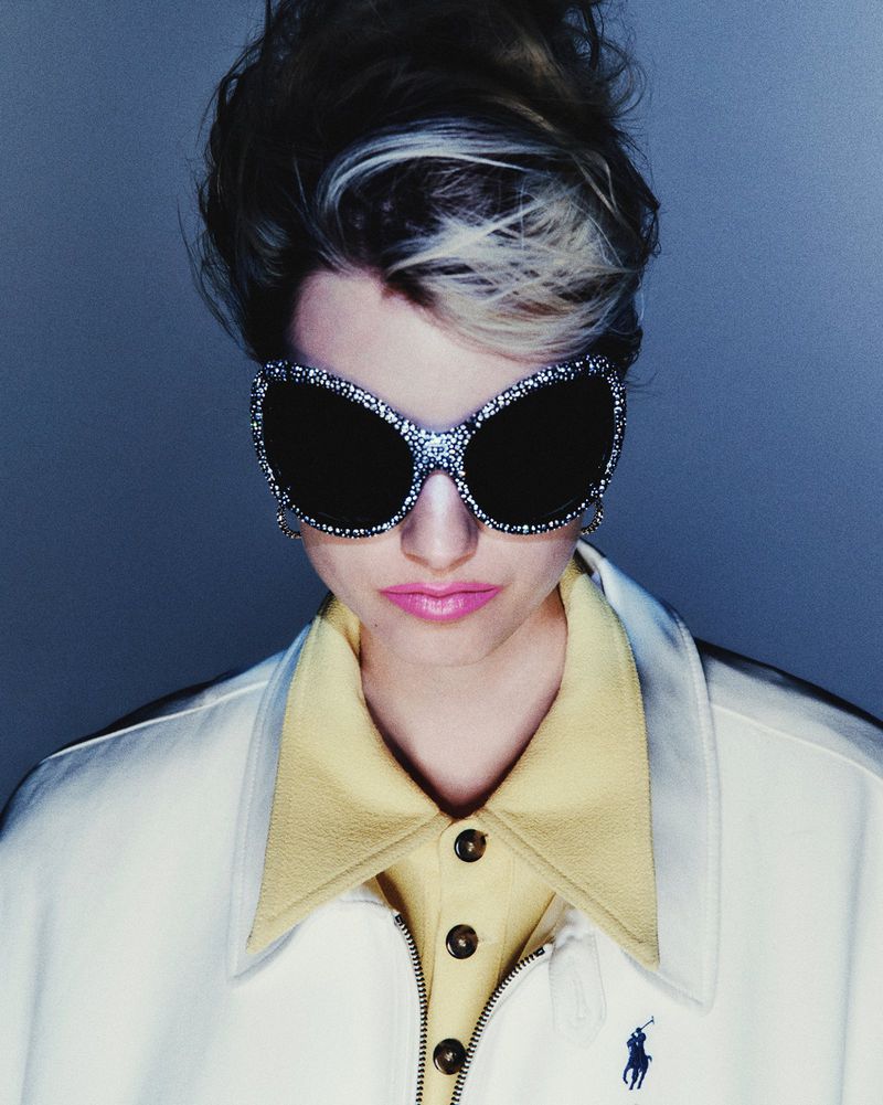 Model Kristin Drab wearing oversized rhinestone sunglasses, a yellow polo shirt, and a white jacket against a grey background.