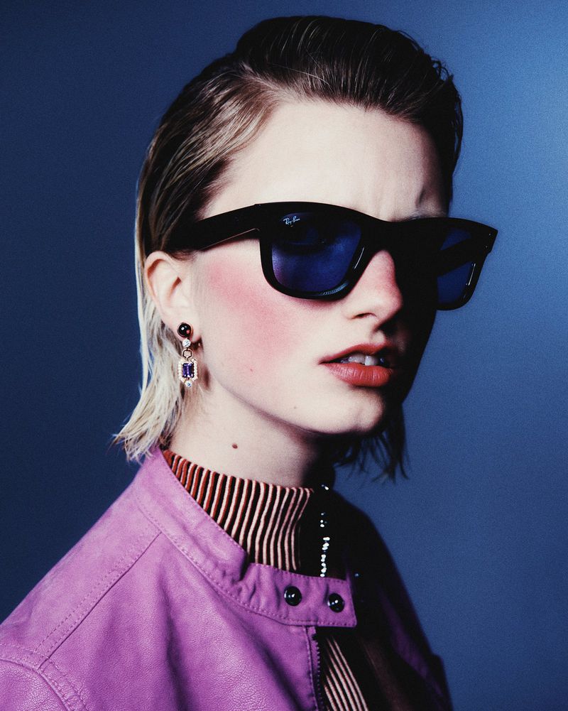 Kristin Drab wearing dark sunglasses, jeweled drop earrings, and a mauve leather jacket against a deep blue background.