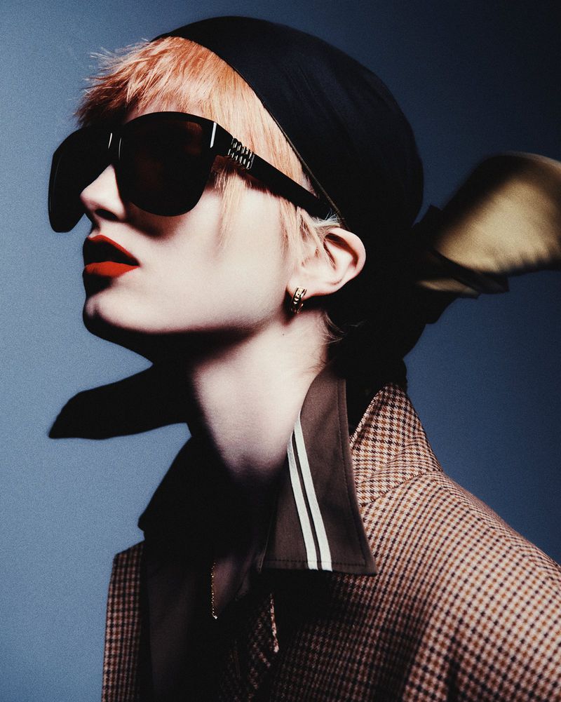 Model Kristin Drab in profile wearing oversized black sunglasses, a black headscarf, and a houndstooth blazer against a blue backdrop.