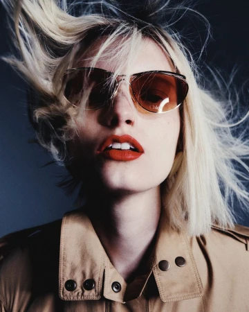 Kristin Drab wearing amber sunglasses, bold red lipstick, and a tan trench coat against a dark grey backdrop.