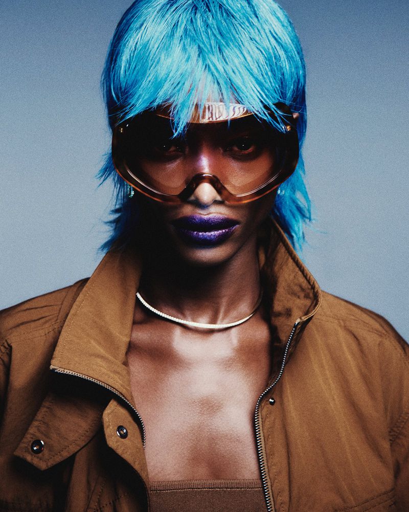 Model Mayowa Nicholas wears an electric blue wig, oversized tinted visor sunglasses, and a brown jacket against a studio backdrop.