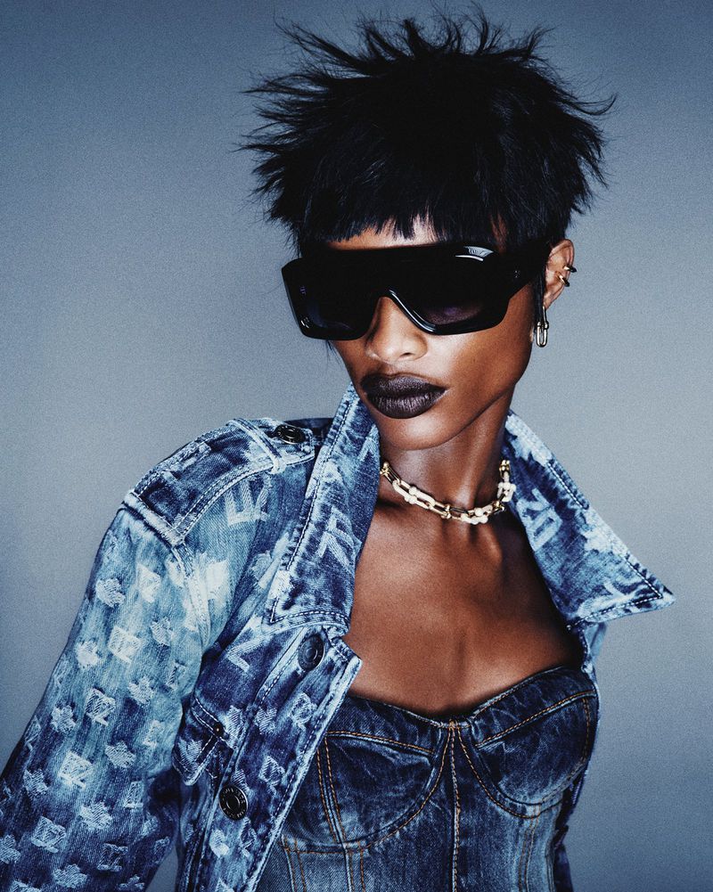 Model Mayowa Nicholas wearing black shield sunglasses and a denim jacket with spiky hair against a blue-gray studio backdrop.