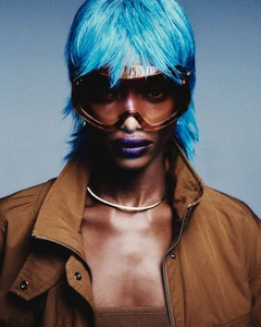 Model Mayowa Nicholas wears an electric blue wig, oversized tinted visor sunglasses, and a brown jacket against a studio backdrop.