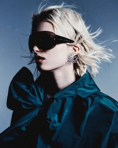 Kristin Drab wearing oversized black shield sunglasses, a crystal earring, and a teal bow collar against a blue background.