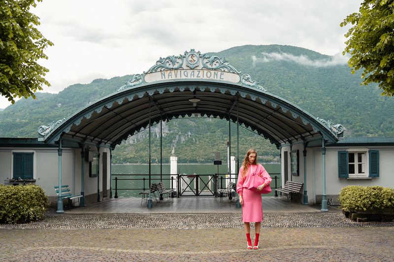 Model Kira Alferink in a pink outfit holding a tote bag stands beneath an ornate ferry arch overlooking a calm mountain lake.