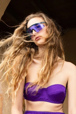 Model Kira Alferink wearing mirrored purple visor sunglasses and a purple strapless top with windswept long blonde hair.