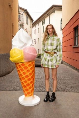 Model Kira Alferink in a green plaid outfit posing next to a giant ice cream cone sculpture in a cobblestone alley.