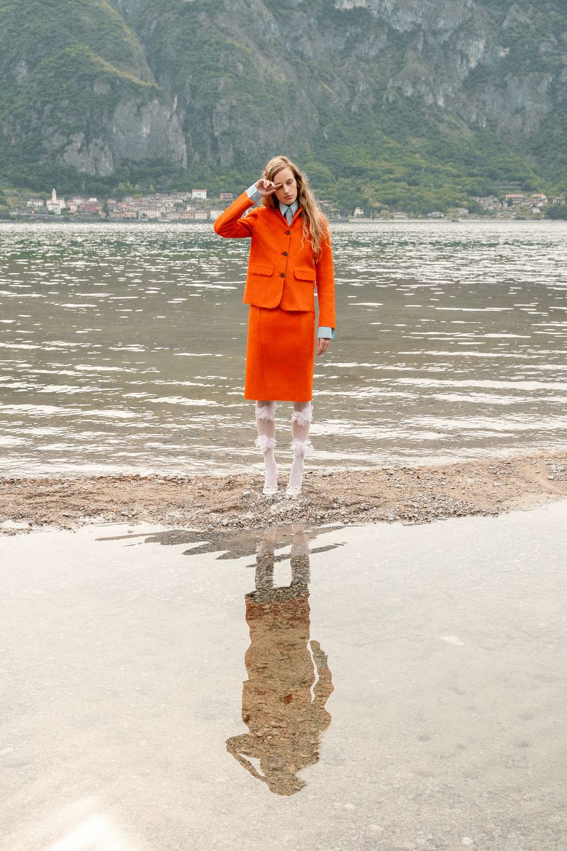 Model Kira Alferink poses in an orange skirt suit by the water with her reflection mirrored in the lake.