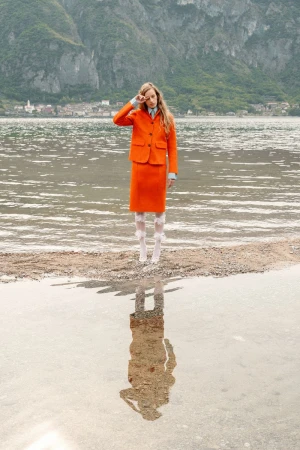 Model Kira Alferink poses in an orange skirt suit by the water with her reflection mirrored in the lake.