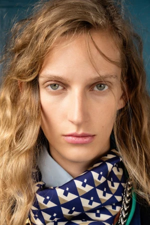 Close-up portrait of model Kira Alferink wearing a geometric patterned blue silk neck scarf and light shirt against a blue backdrop.