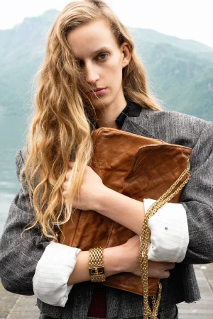 Model with wavy blonde hair clutching a brown quilted leather handbag with gold chain strap wearing a grey tweed jacket outdoors.
