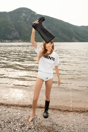 Model Kira Alferink pours water from a black boot over her head on a pebbled lakeshore wearing a white t-shirt.