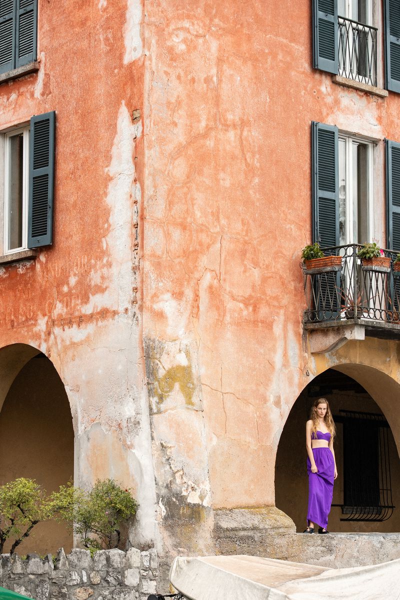 Model Kira Alferink in a vibrant purple two-piece dress standing beneath an arched portico of a weathered terracotta building.
