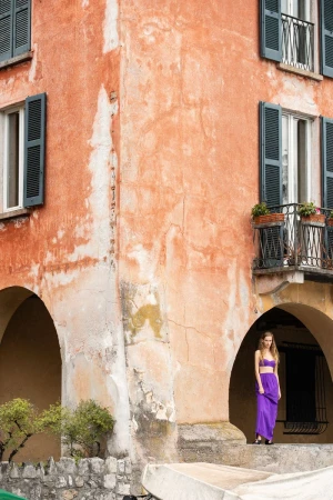 Model Kira Alferink in a vibrant purple two-piece dress standing beneath an arched portico of a weathered terracotta building.