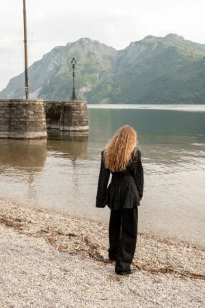 Model Kira Alferink viewed from behind in a black textured jacket and trousers on a pebbly lake shoreline near mountains.