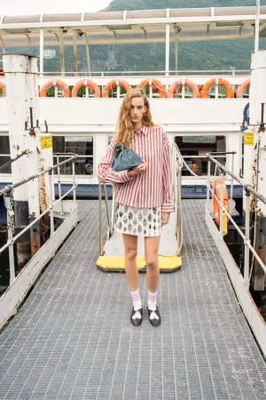 Model Kira Alferink standing on a boat gangway holding a blue handbag in a pink striped shirt and silver patterned mini skirt.