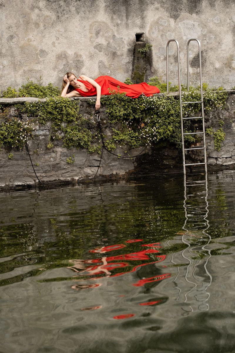 Woman in red dress rests on a stone wall covered in greenery, reflected in the water. Ladder leads to water.