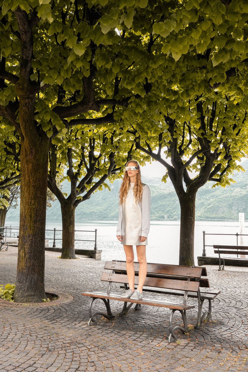 Model Kira Alferink standing on a park bench wearing a white dress and futuristic silver visor under a tree canopy by a lake.