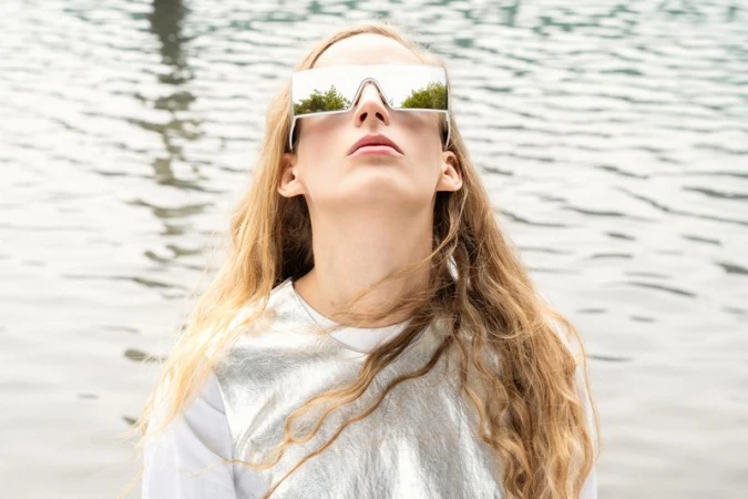 Model Kira Alferink wearing mirrored visor sunglasses and a silver metallic top outdoors against a calm water surface.