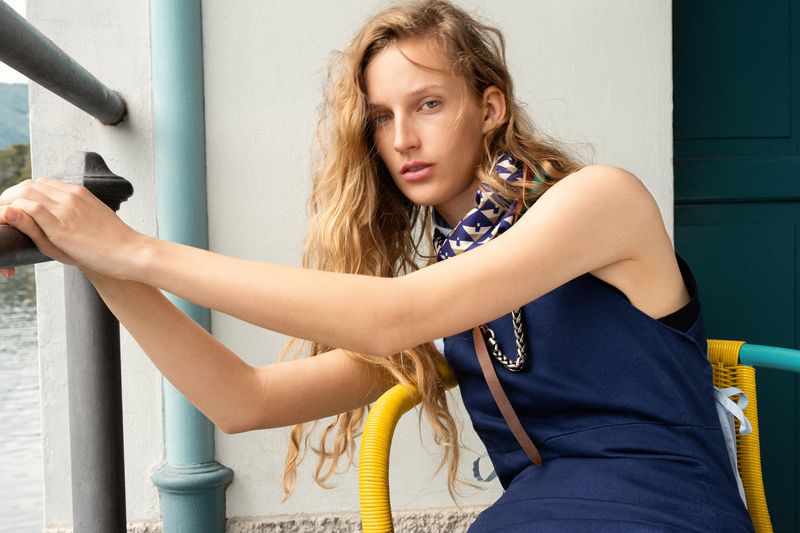 Model Kira Alferink wearing a navy blue sleeveless dress and patterned neck scarf seated near a lakeside railing.