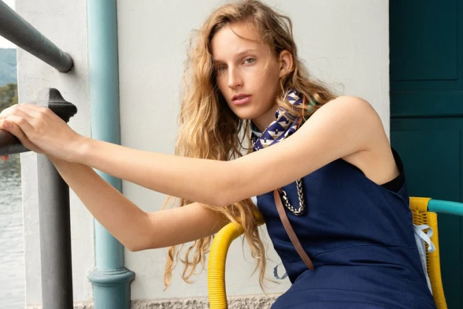 Model Kira Alferink wearing a navy blue sleeveless dress and patterned neck scarf seated near a lakeside railing.