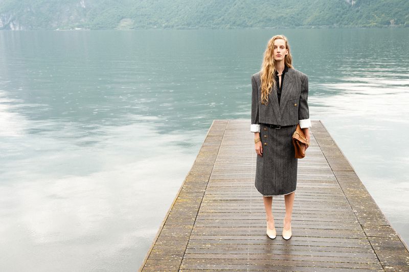 Model standing on a wooden pier overlooking a lake wearing a grey tweed suit jacket and matching skirt holding a brown leather clutch
