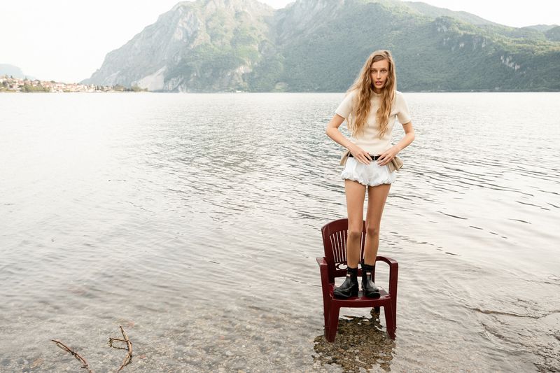 Model Kira Alferink standing on a plastic chair in shallow lake water wearing a cream top, white bloomer shorts, and black boots.