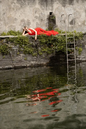 Model Kira Alferink reclining in a red dress on a stone ledge over reflective water beside a metal pool ladder.