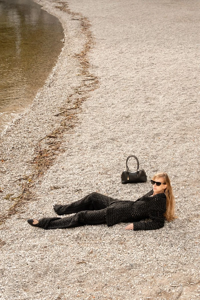 Model Kira Alferink in a black textured knit suit and sunglasses lying on a pebbled beach beside a black handbag.
