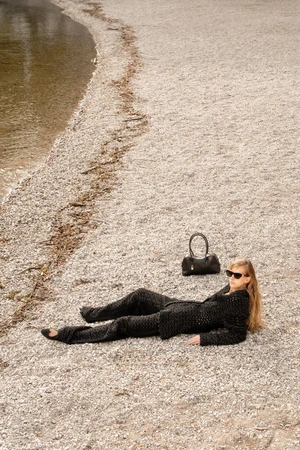 Model Kira Alferink in a black textured knit suit and sunglasses lying on a pebbled beach beside a black handbag.